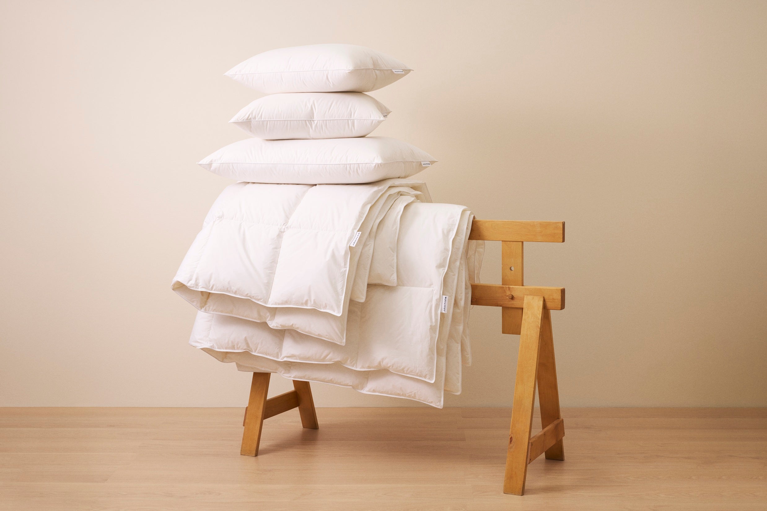 Stack of white pillows and a blanket on a wooden stool against a beige wall.