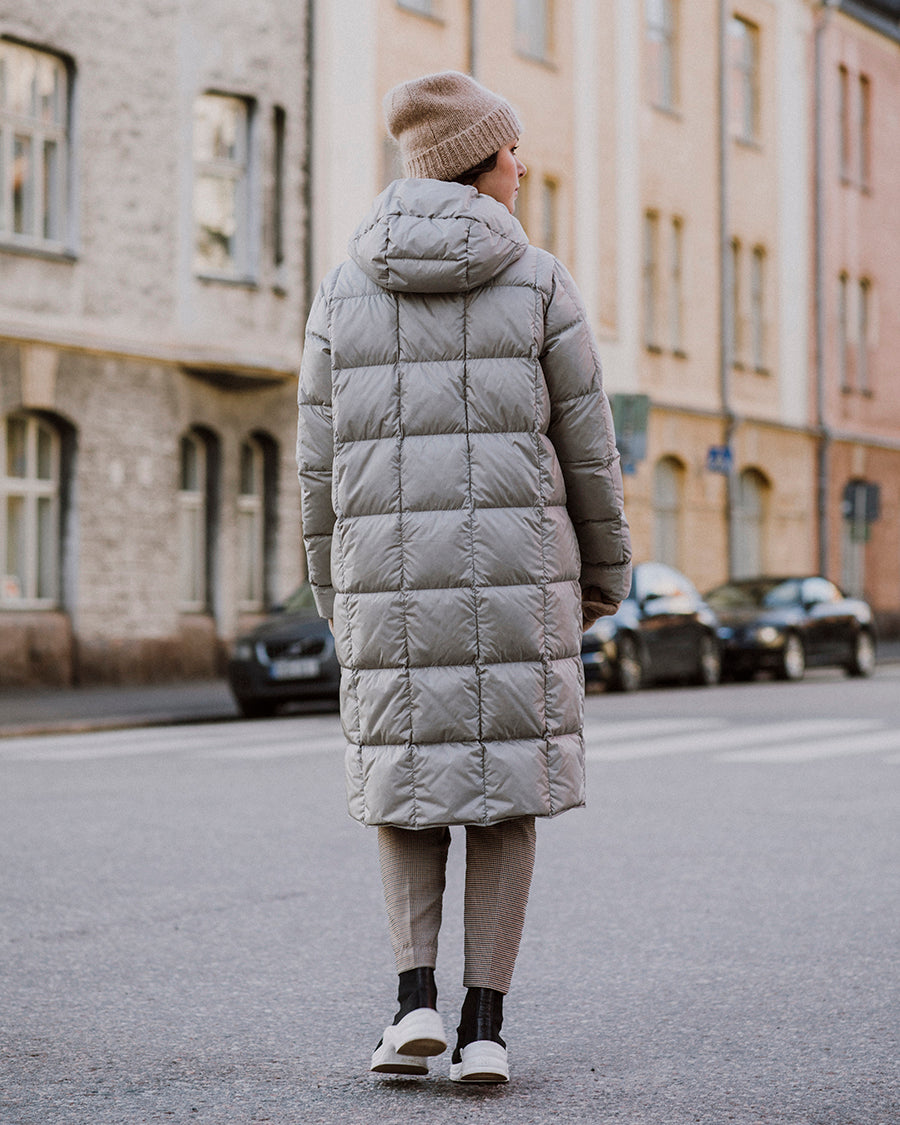 A woman on the street with her back to the camera, wearing a light, long Essi light down coat (color: champagne) with a hood and square quilting. Her outfit includes a beige beanie, striped pants, and black-and-white shoes. Apartment buildings and parked cars are visible in the background.