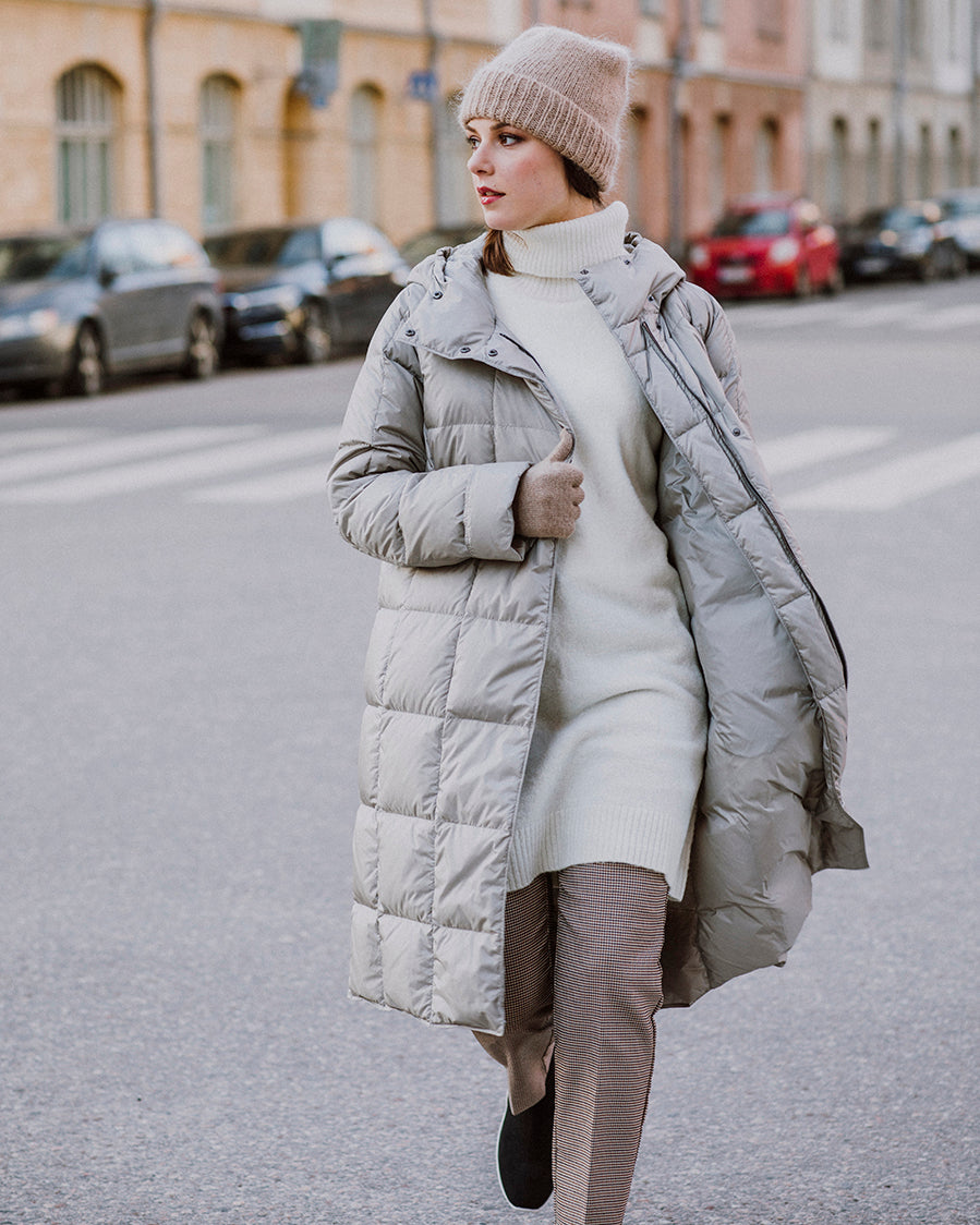 A woman walks down the street wearing a light, long Essi light down coat (color: champagne) with square quilting, worn open. Her outfit also includes a beige beanie, a light knit dress, striped pants, and beige gloves. In the background, there is an urban cityscape with parked cars.