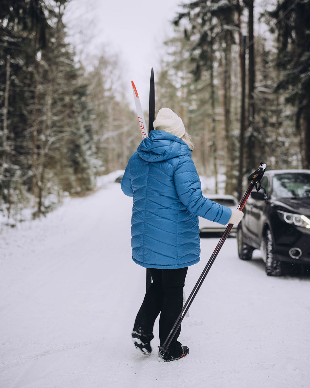 A woman is walking on a snowy road holding skis and poles. She is wearing a light blue, quilted Jasmin light down coat (color: Blue butterfly) with a hood, and a white beanie.
