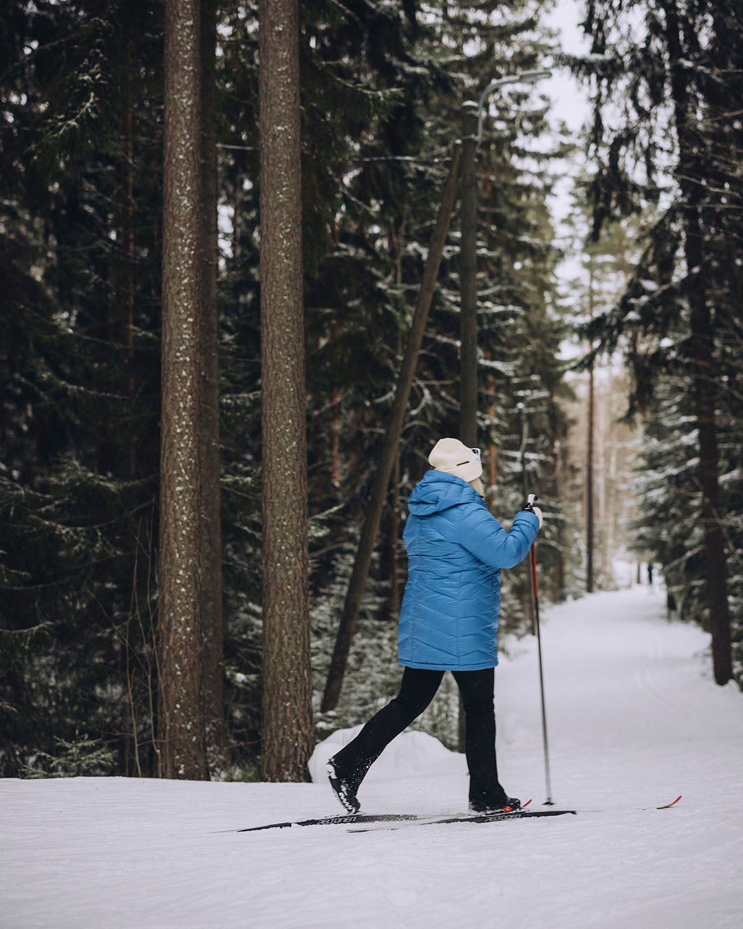 A woman is skiing in a snowy forest. She is wearing a light blue Jasmin light down coat (color: Blue butterfly), black trousers, and a white beanie.