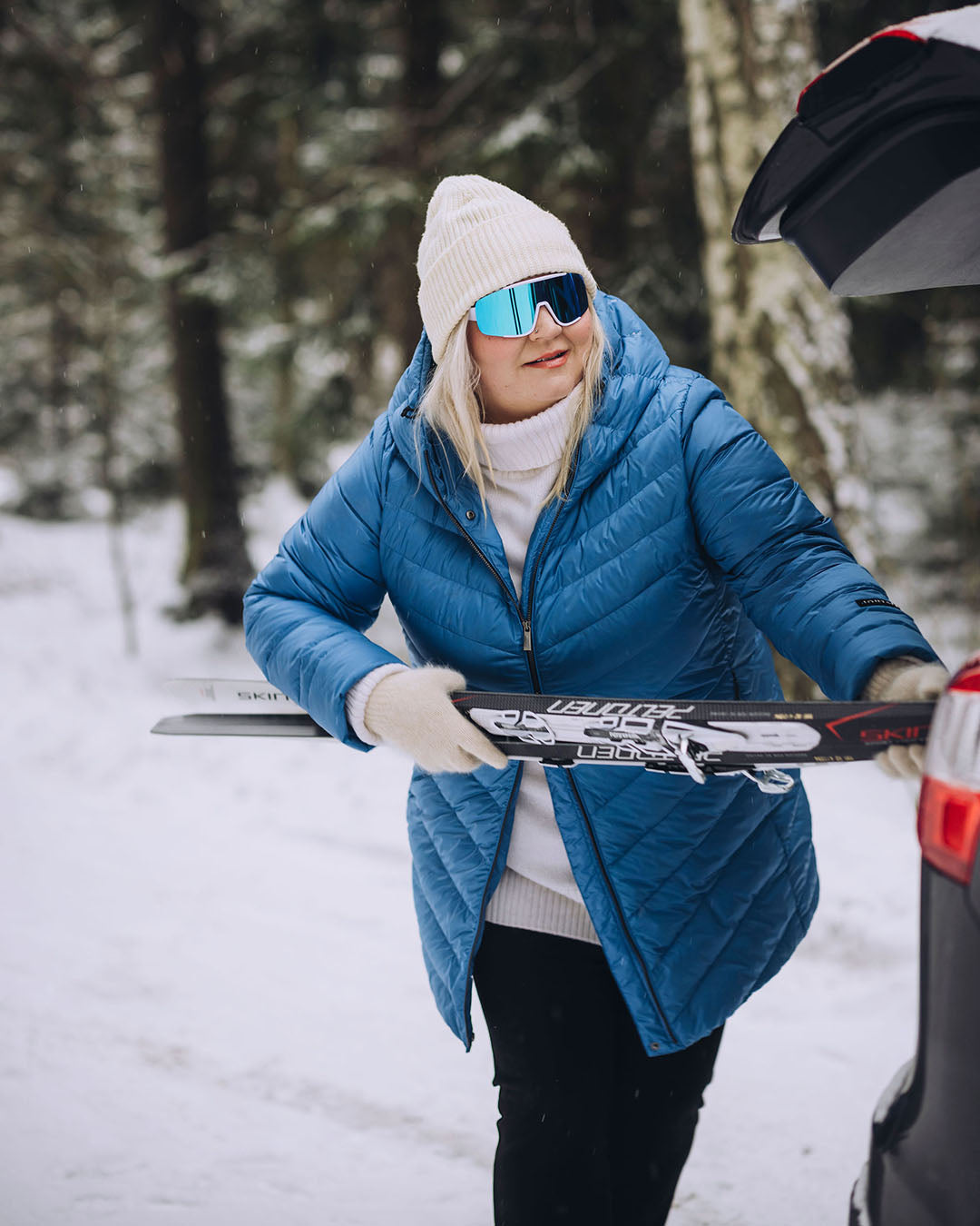 A woman is standing in a snowy forest, taking skis out of a car trunk. She is wearing a light blue Jasmin light down coats (color: Blue butterfly), a white beanie, and reflective ski goggles.