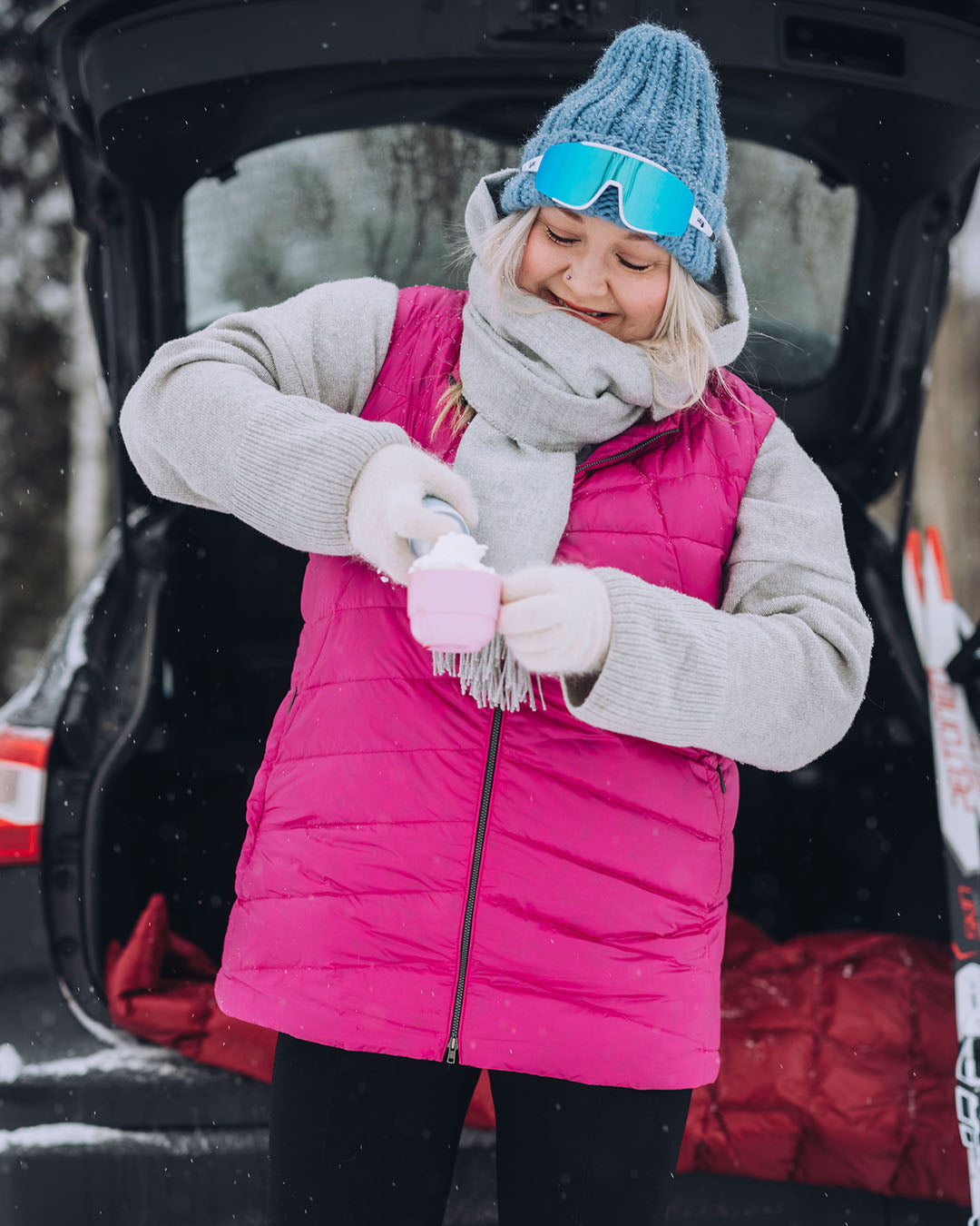A woman is standing in front of an open car trunk, pouring a drink into a cup. She is wearing a pink Mesi down vest (color: Pitaya), a grey knit sweater, and a blue beanie, with ski goggles resting on her forehead.