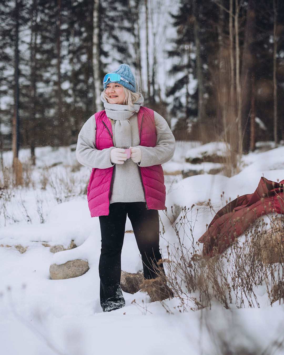 A woman is standing in a snowy forest landscape, wearing a pink Mesi down vest (color: Pitaya), a grey knit sweater, and black trousers. She has a blue beanie and sunglasses, and she is smiling while looking to the side.
