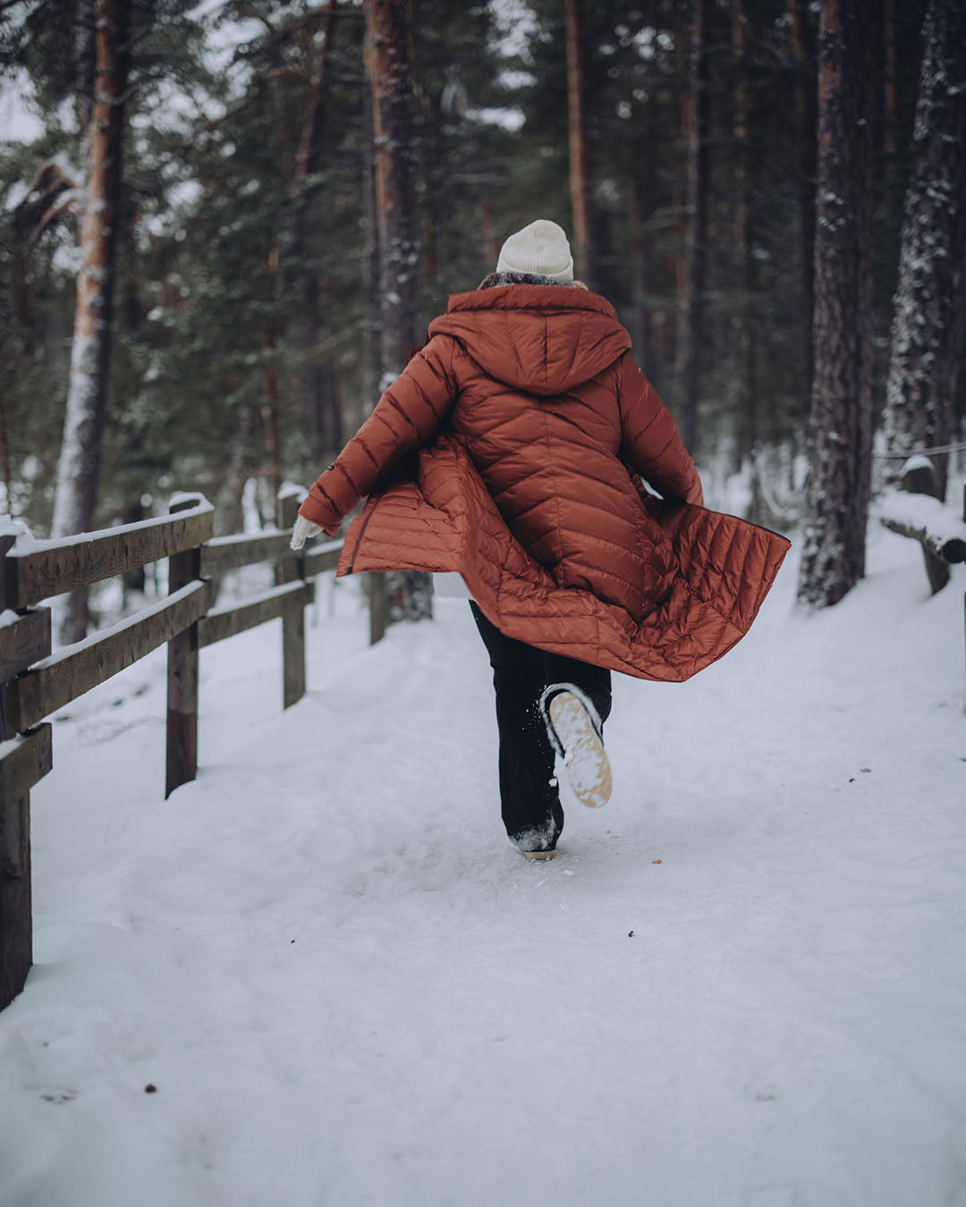 A woman is running with her coat open on a snowy forest trail, wearing a muted orange, quilted Säde light down coat (color: rose gold) and a white beanie. There is a fence to her left.