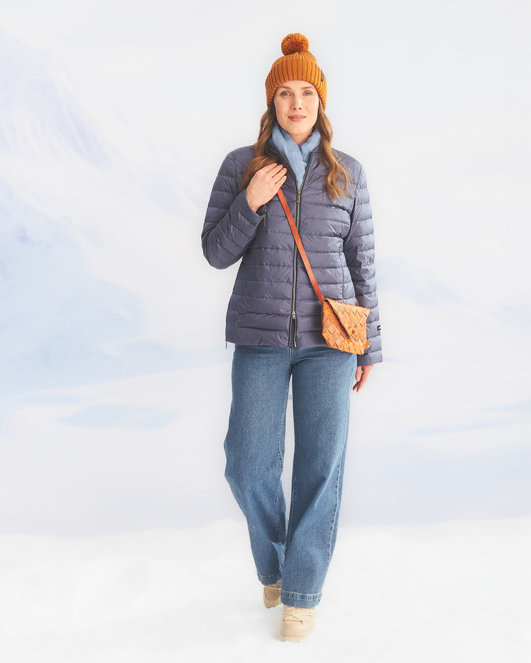 A woman stands in a snowy landscape wearing a blue Maana light down coat (color: Storm), a blue scarf, and an orange pom-pom beanie. Her outfit also includes wide-leg blue jeans and a brown woven shoulder bag.