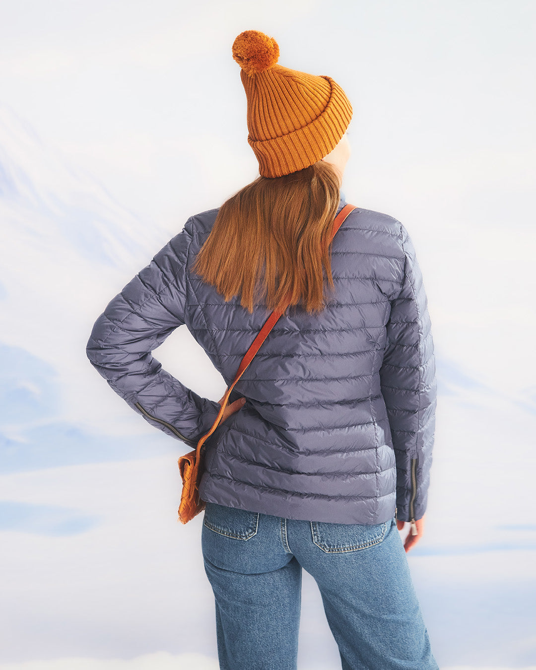 A woman stands with her back to the camera in a snowy landscape, wearing a blue Maana light down coat (color: Storm), blue jeans, and an orange pom-pom beanie. She carries a brown woven shoulder bag on her shoulder.