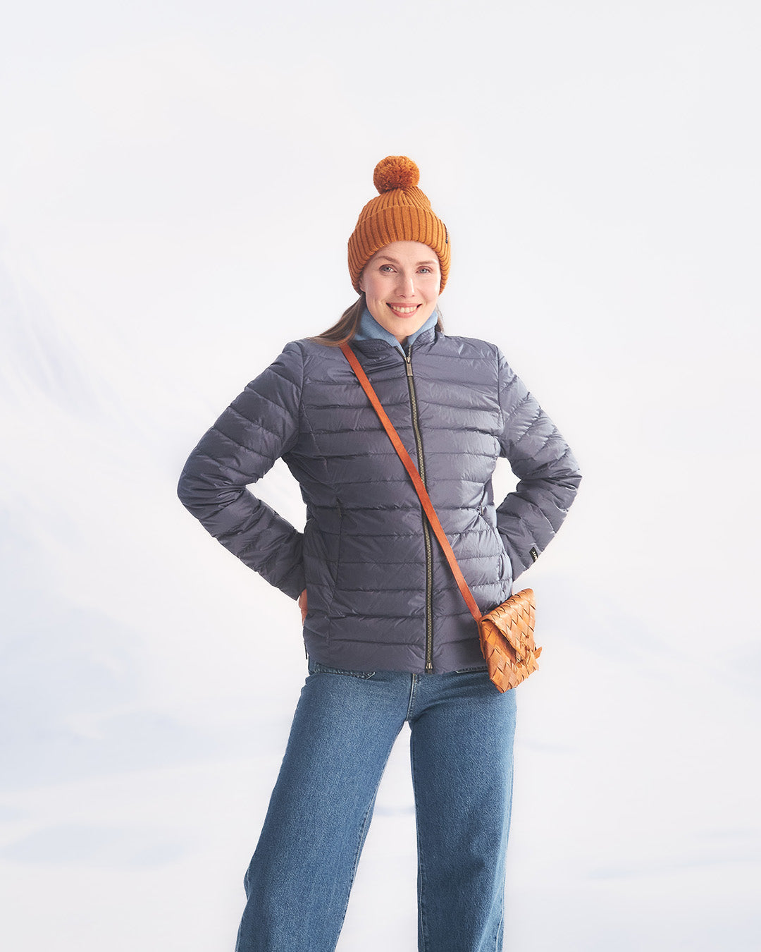 A smiling woman stands in a snowy setting wearing a blue Maana light down coat (color: Storm), blue jeans, and an orange pom-pom beanie. She carries a brown woven shoulder bag.