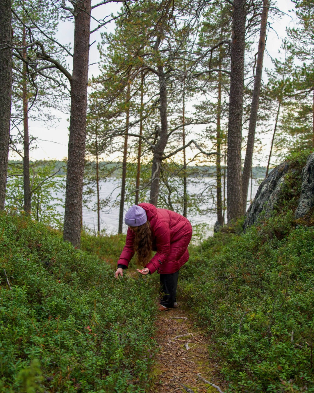 Person in a dark red light down coat and a lilac beanie walking through a forest with a lake in the background