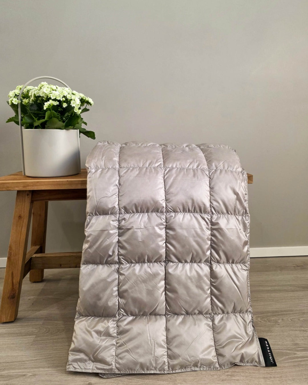 Gray Kulkuri down blanket on a wooden stool with a plant against a plain wall.