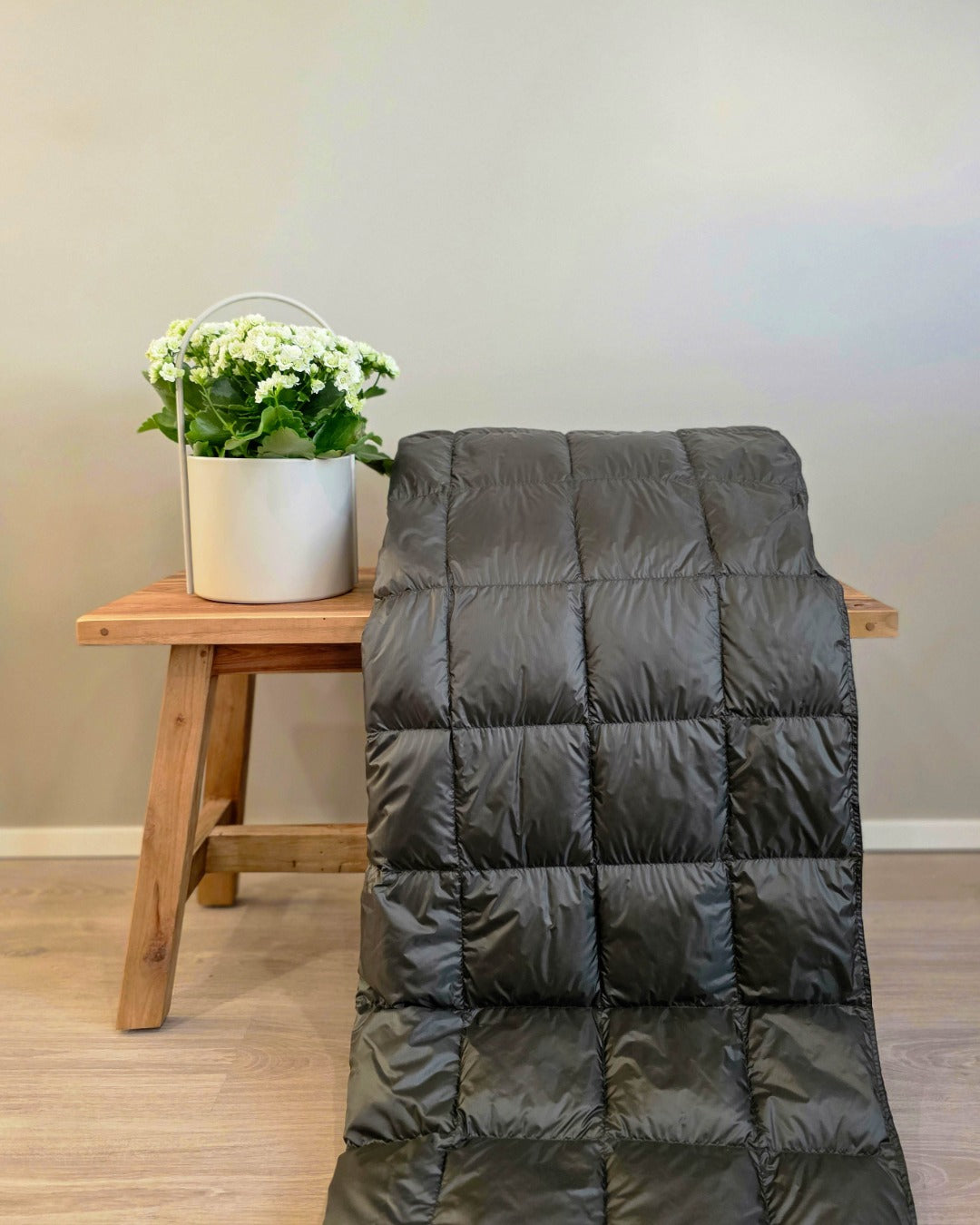 Dark green quilted Kulkuri down blanket draped over a chair next to a wooden stool with a plant on a light wood floor.