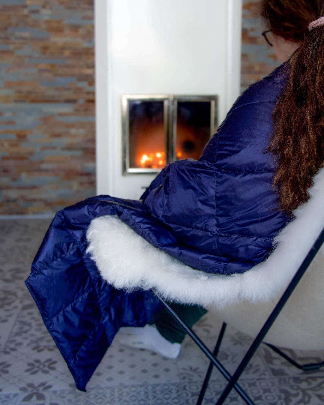 Person wearing a dark blue Kulkuri down blanker on her sholders with white fur trim sitting in front of a fireplace.