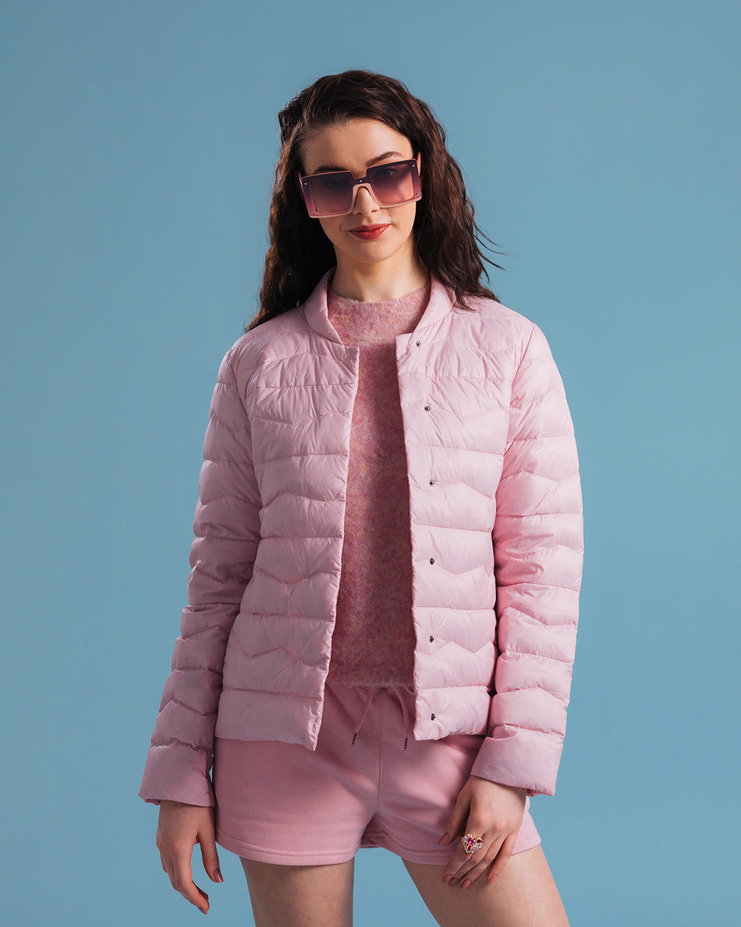 A woman wearing a light pink Lootus light down jacket (color: strawberry mousse), a matching fluffy knit sweater, and shorts. The outfit is completed with oversized sunglasses and a ring.
