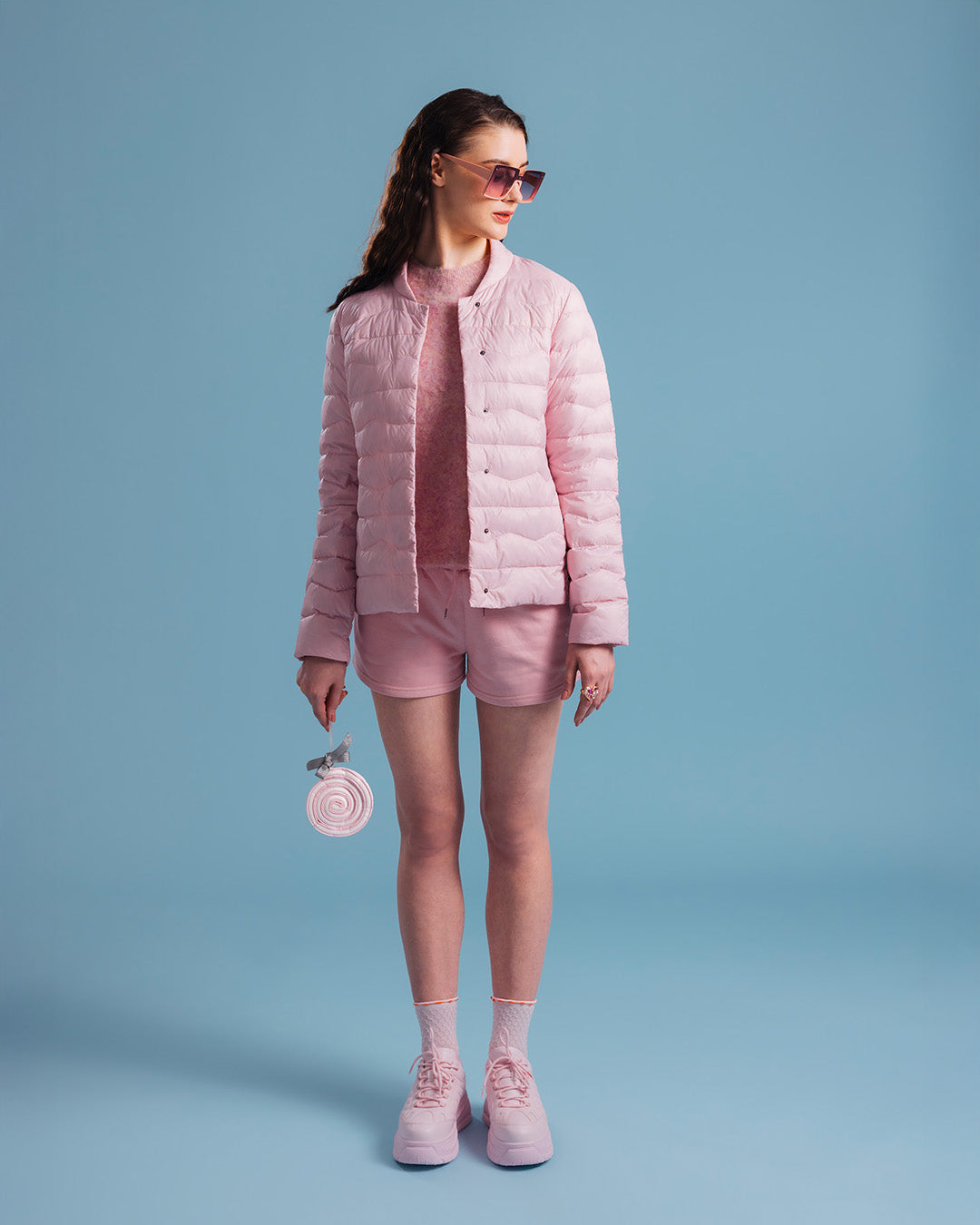 A woman dressed in a light pink Lootus light down jacket (color: strawberry mousse), a matching knit sweater and shorts. Her outfit includes pink sneakers, sunglasses, and a large round lollipop in her hand.