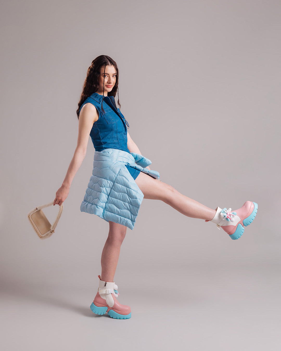 A woman wearing a sleeveless denim dress, with a light blue Lootus light down jacket (color: Frozen candy) tied around her waist, and light pink shoes. The outfit is completed with a beige handbag.