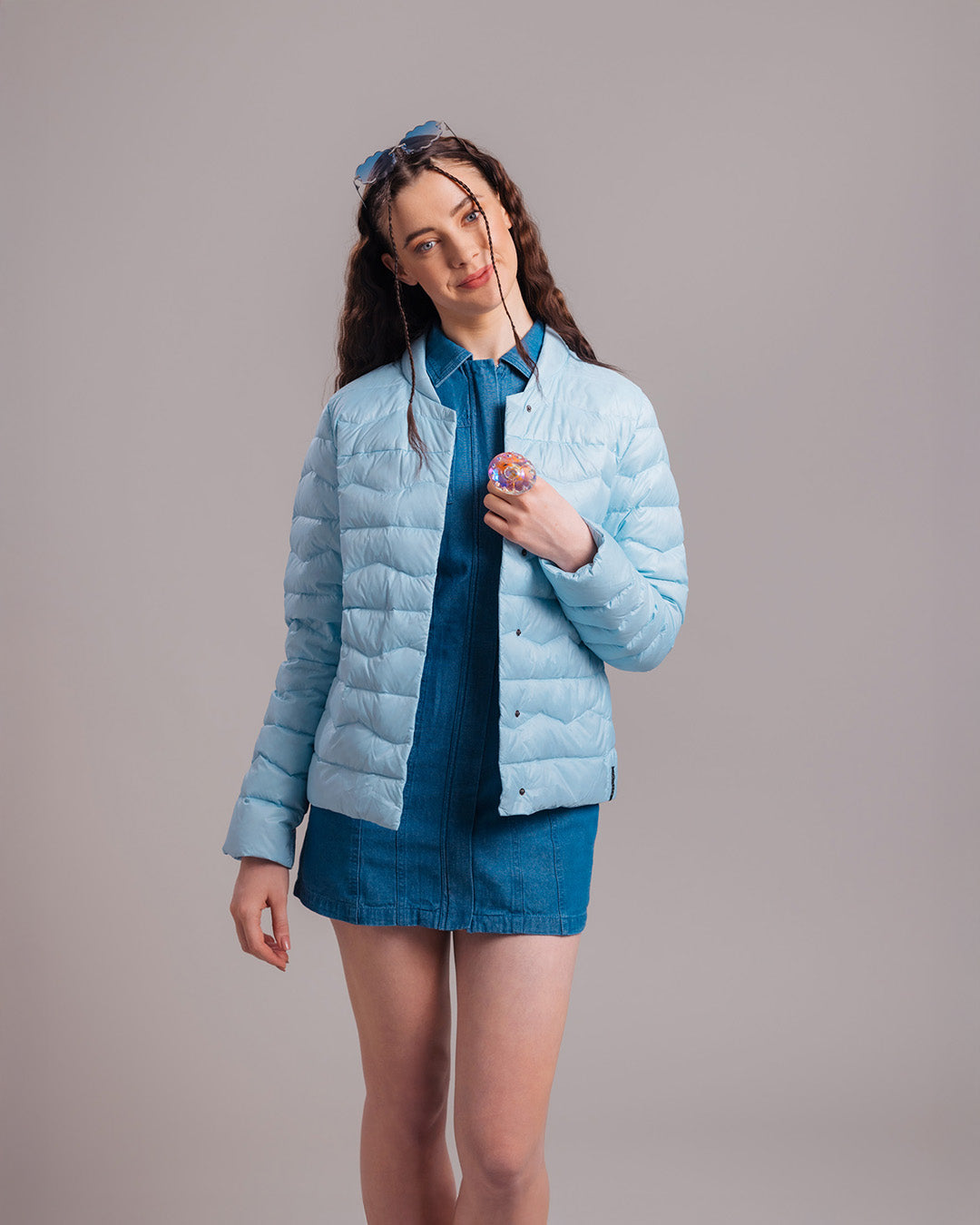 A woman wearing a light blue Lootus light down jacket (color: Frozen candy) and a blue denim dress. The outfit is complemented by sunglasses worn in her hair and a large statement ring.
