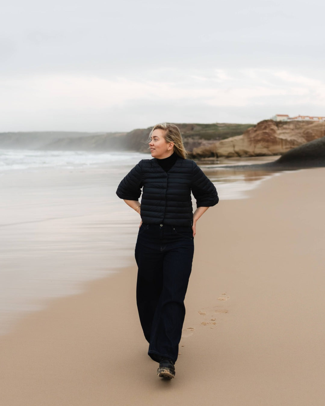 Person in black down bolero walking on a beach with ocean and cliffs in the background