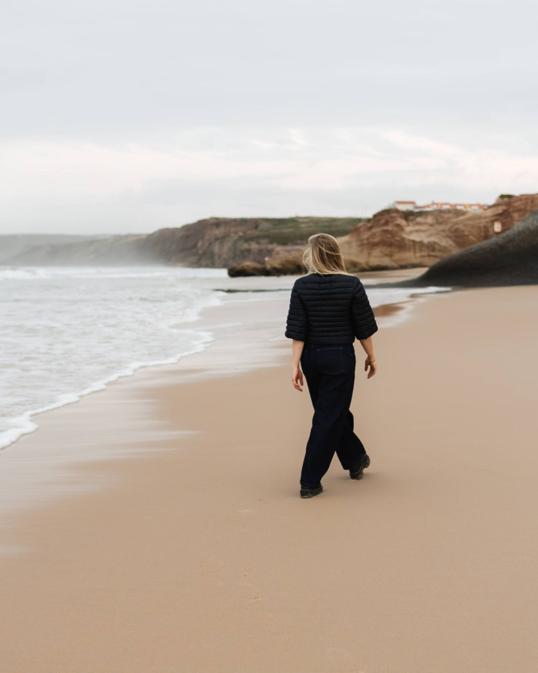 Person walking on a beach with cliffs in the background. She is wearing a black down bolero.