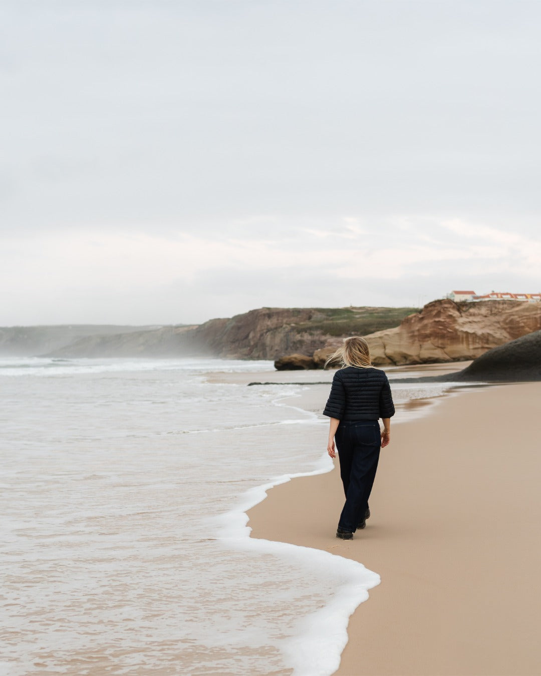 Person walking on a beach with cliffs and ocean waves in the background. She is wearing a black down bolero