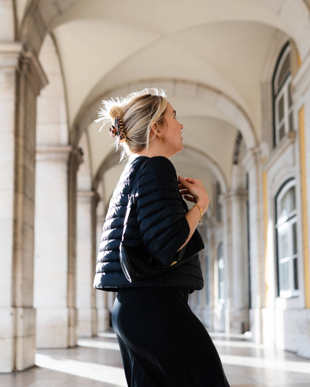 Woman in a black down bolero standing in an archway with classical architecture.