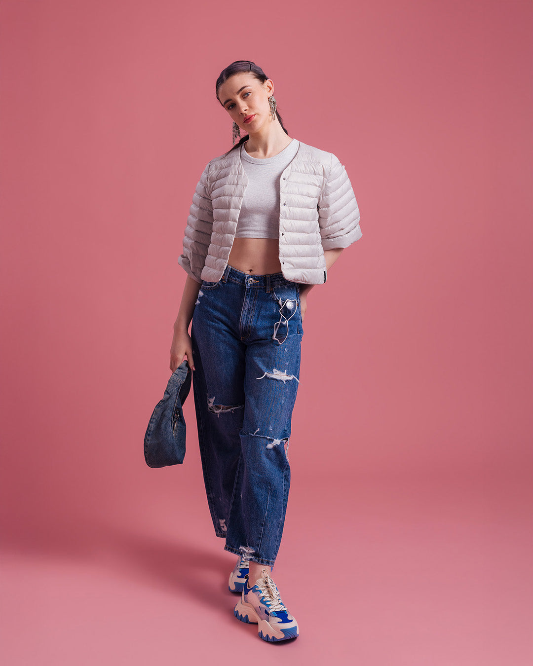 A woman dressed in a light-colored, short Lupiini light down jacket (color: sugar cloud), a light short top, and ripped blue jeans. The outfit is accessorized with earrings, blue sneakers, and a dark handbag.