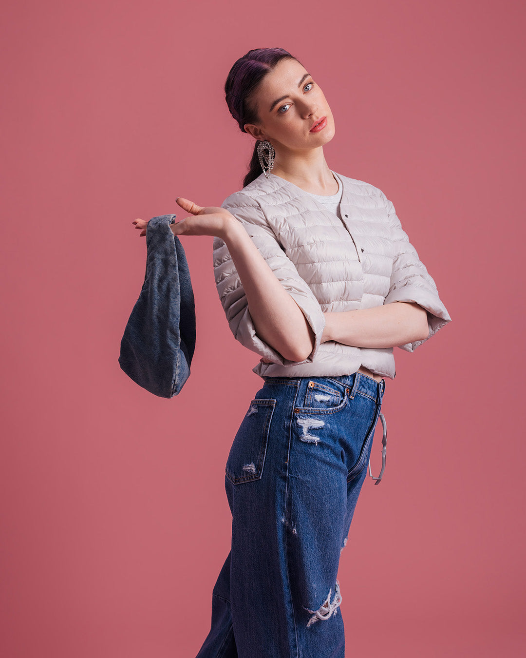 A woman stands sideways wearing a light-colored, short Lupiini lightweight down jacket (color: sugar cloud) and ripped blue jeans. She holds a dark handbag in one hand.