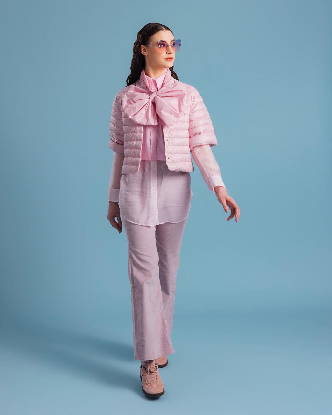 A woman wearing a light pink, short Lupiini light down jacket (color: strawberry mousse) adorned with a striking bow detail on top, layered over a sheer long collared shirt. The outfit includes light-colored trousers, pink shoes, and purple sunglasses.