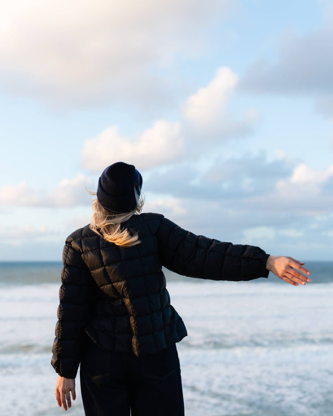 Person standing on a beach with arms outstretched, facing the ocean. She is vearing a black down college