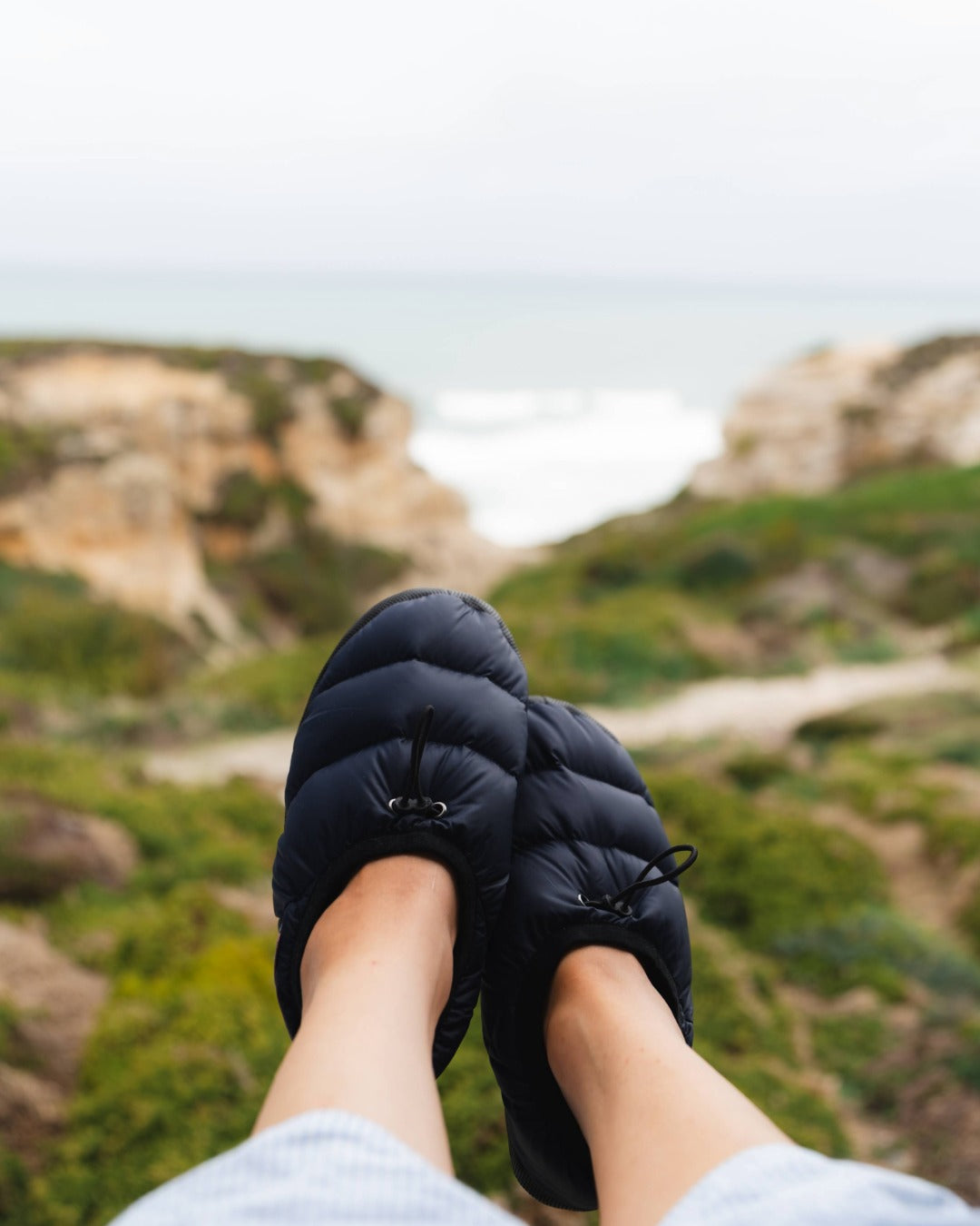 Person wearing black camp down slippers with a scenic background of green hills and mountains.