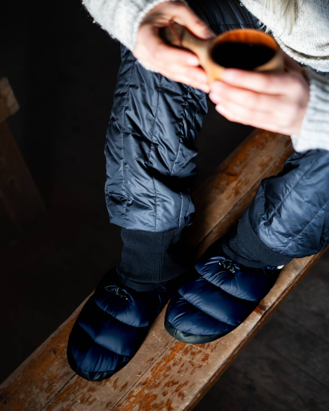 Person wearing blue camp down slippers sitting on a wooden bench holding a small cup.
