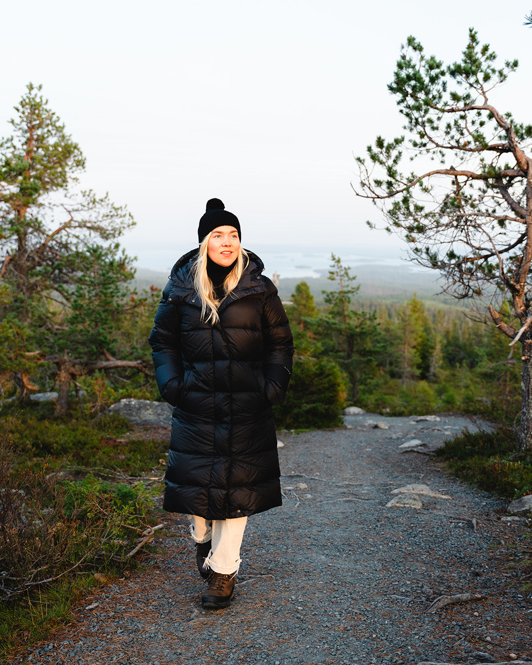A person walks on a rocky path in a forested landscape wearing a long, black (color name: charcoal black) Nietos down coat. The coat reaches the calves and has a quilted surface, high collar, and attached hood. The person also wears a black beanie, light-colored pants, and sturdy hiking shoes. In the background, there is a pine forest and a distant view of water and trees in the evening light.