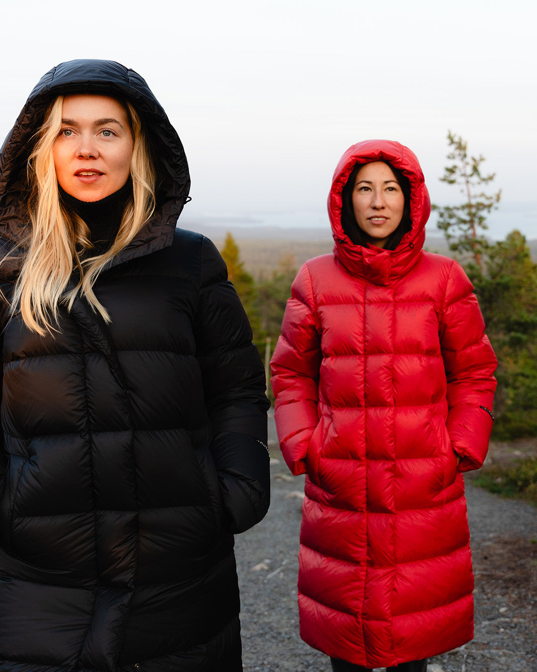Two women are standing on a rock with trees visible in the background. The blonde woman is wearing a black (color: charcoal black) down coat, and the dark-haired woman is wearing a long red (color: raspberry) down coat. Both have their hoods up and hands in their coat pockets. They are looking past the camera with neutral expressions on their faces.