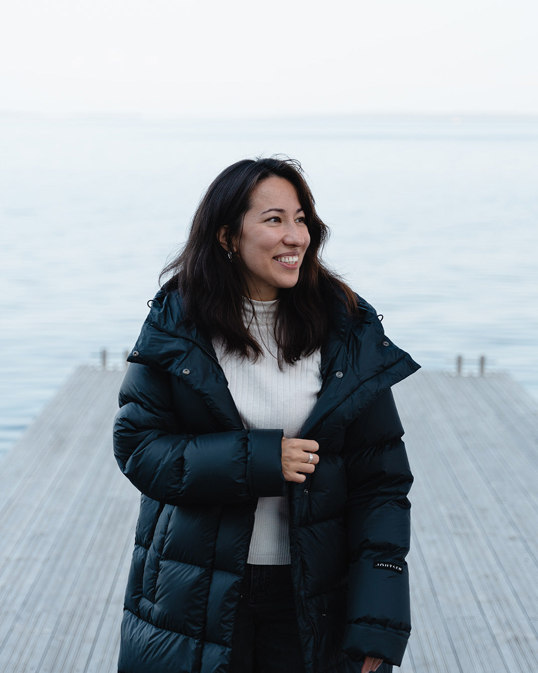 A woman stands on a large pier with a foggy body of water in the background. She is wearing a black (color: charcoal black) down coat with the zipper open and is holding the coat’s edge with one hand.