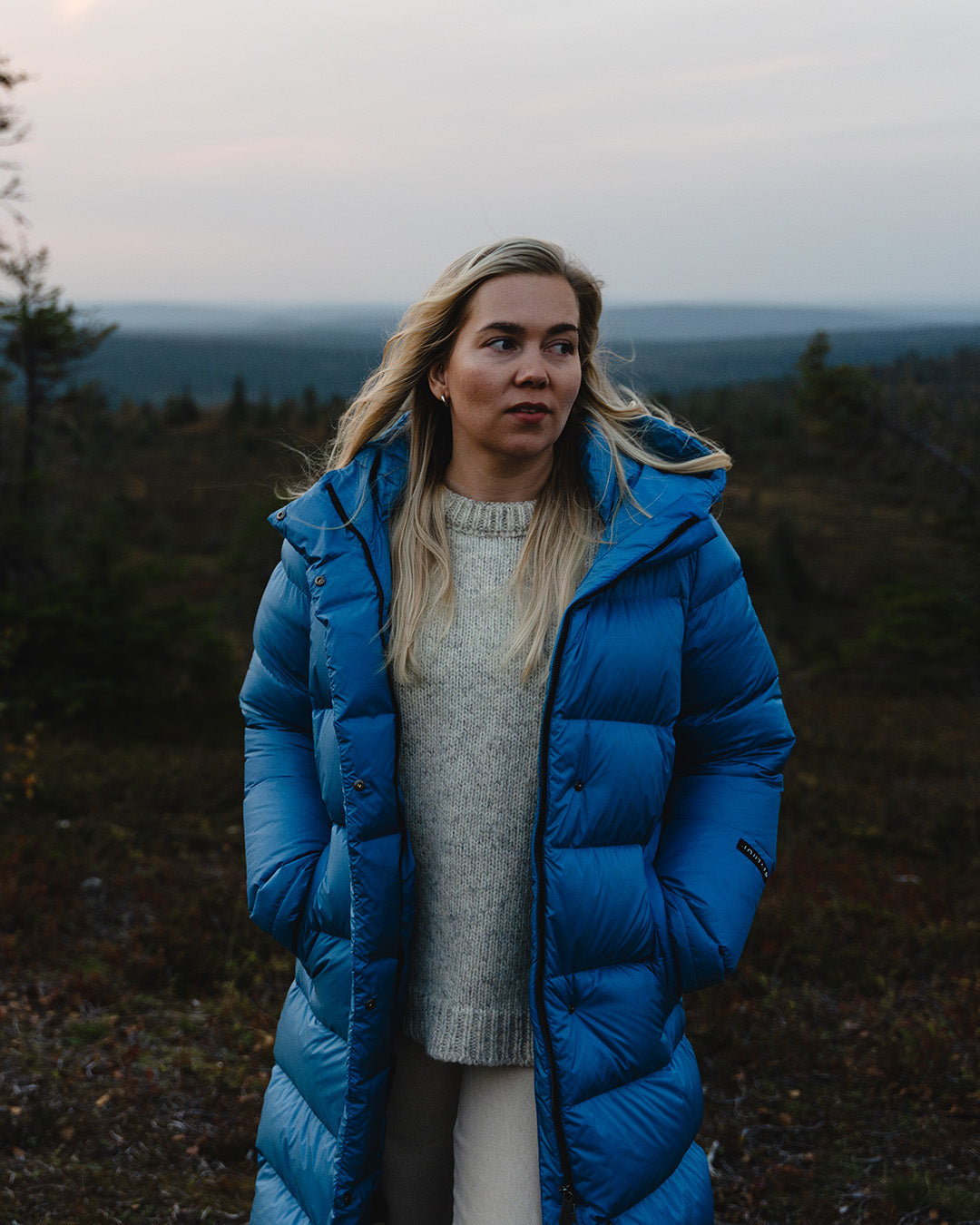 A blonde woman stands in a forested landscape. She is wearing a long blue (color: Blue butterfly) down coat with the zipper open and her hands in the coat pockets. Underneath, she has light-colored pants and a light sweater.