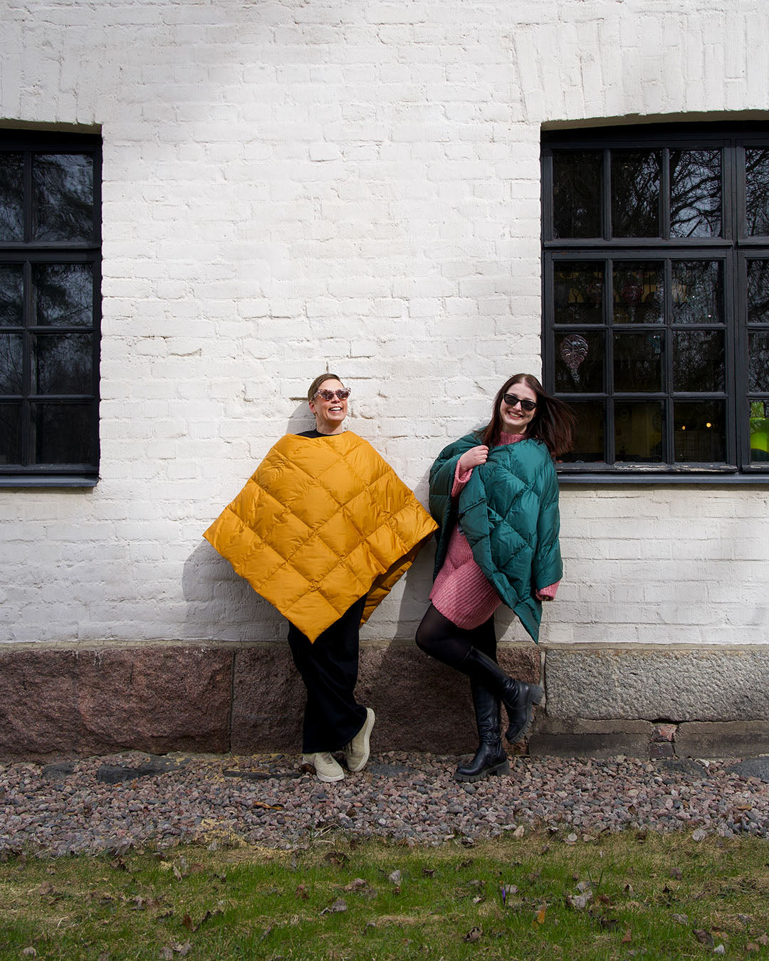 Two people standing outdoors in front of a white brick wall, wearing loose Otava down ponchos with diamond quilting. The person on the left wears a yellow (amber) poncho, and the person on the right wears a dark green (green paprika) poncho. Both wear sunglasses and smile.