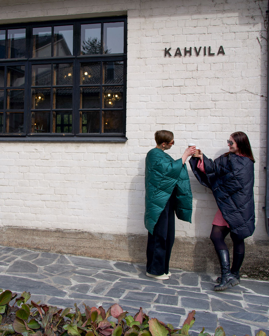 Two people standing in front of a café’s white brick wall, clinking drink mugs. Both wear Otava down ponchos with diamond quilting—one green (green paprika) and the other charcoal black. They are wearing sunglasses.