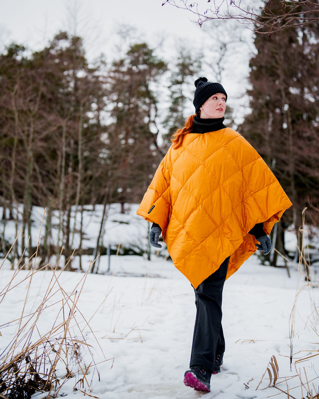 Person walking in a snowy forest wearing a yellow Otava down poncho (color: amber), black beanie, gray gloves, and dark pants. The poncho features a diamond-shaped quilting pattern.