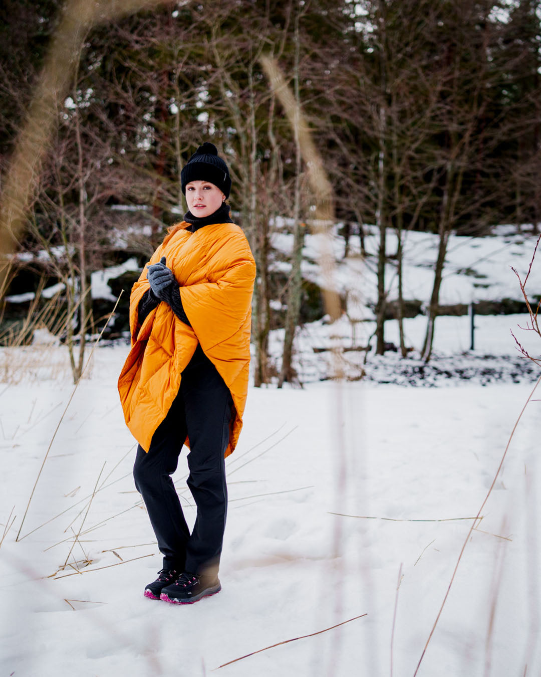 Person standing in a snowy forest wearing a yellow Otava down poncho (color: amber), black beanie, gray gloves, and dark pants. Snow covers the ground and trees are visible in the background.