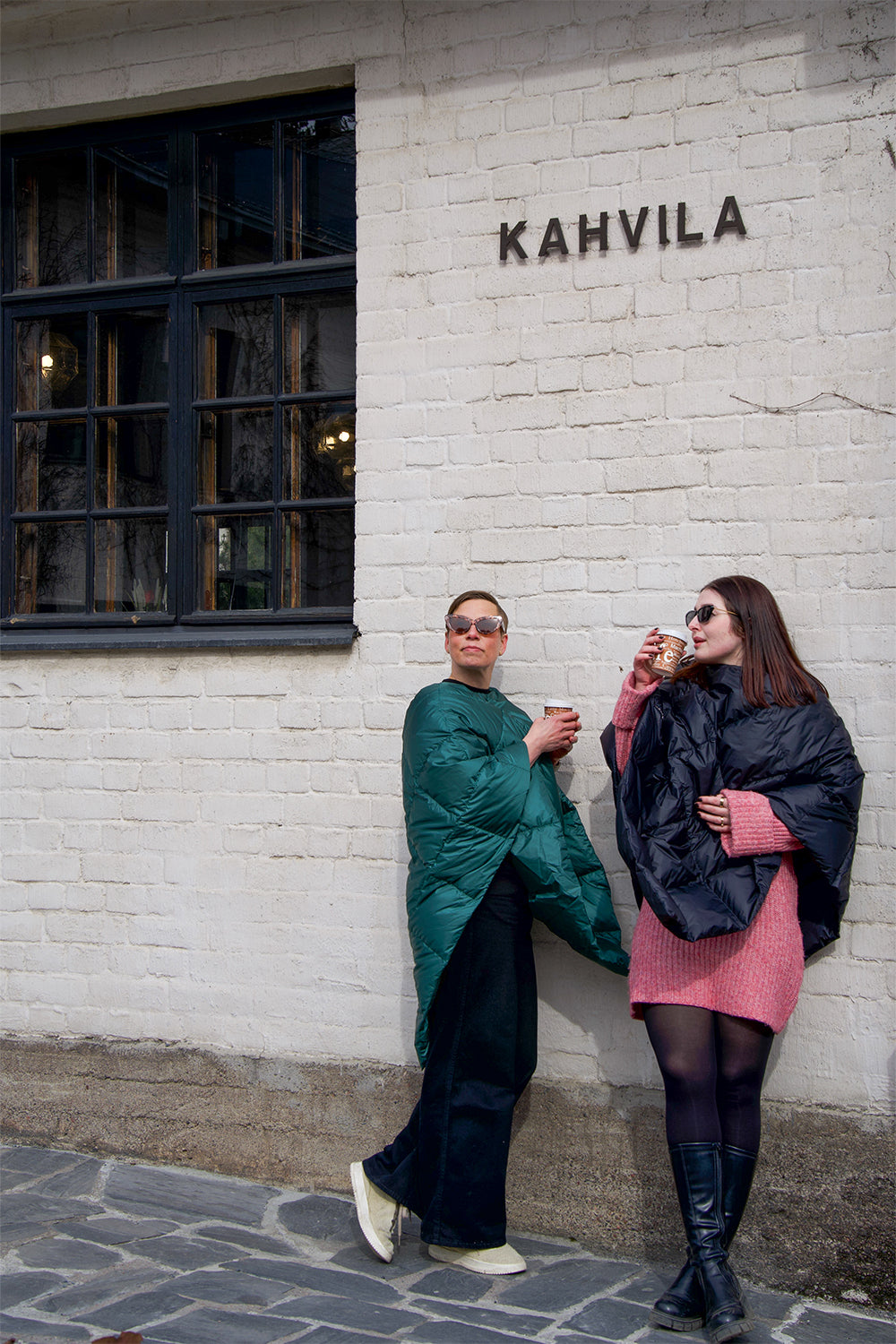 Two people are standing in front of a café’s white brick wall, drinking coffee. Both are wearing diamond-quilted Otava down ponchos—one in green (color: green paprika) and the other in charcoal black. They are wearing sunglasses.
