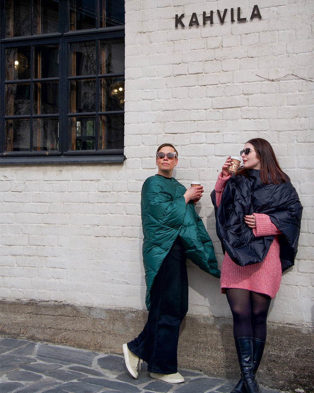 Two people are standing in front of a café’s white brick wall, drinking coffee. Both are wearing diamond-quilted Otava down ponchos—one in green (color: green paprika) and the other in charcoal black. They are wearing sunglasses.