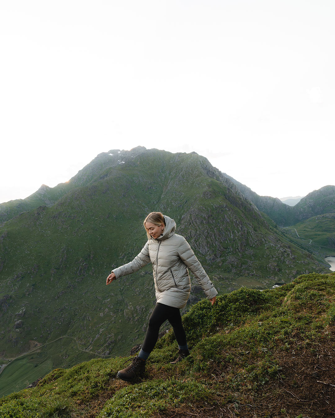 A woman walks down a slope wearing a beige Ruut light down coat (color: champagne), black leggings, and hiking boots. In the background, a lush mountainous landscape is visible.