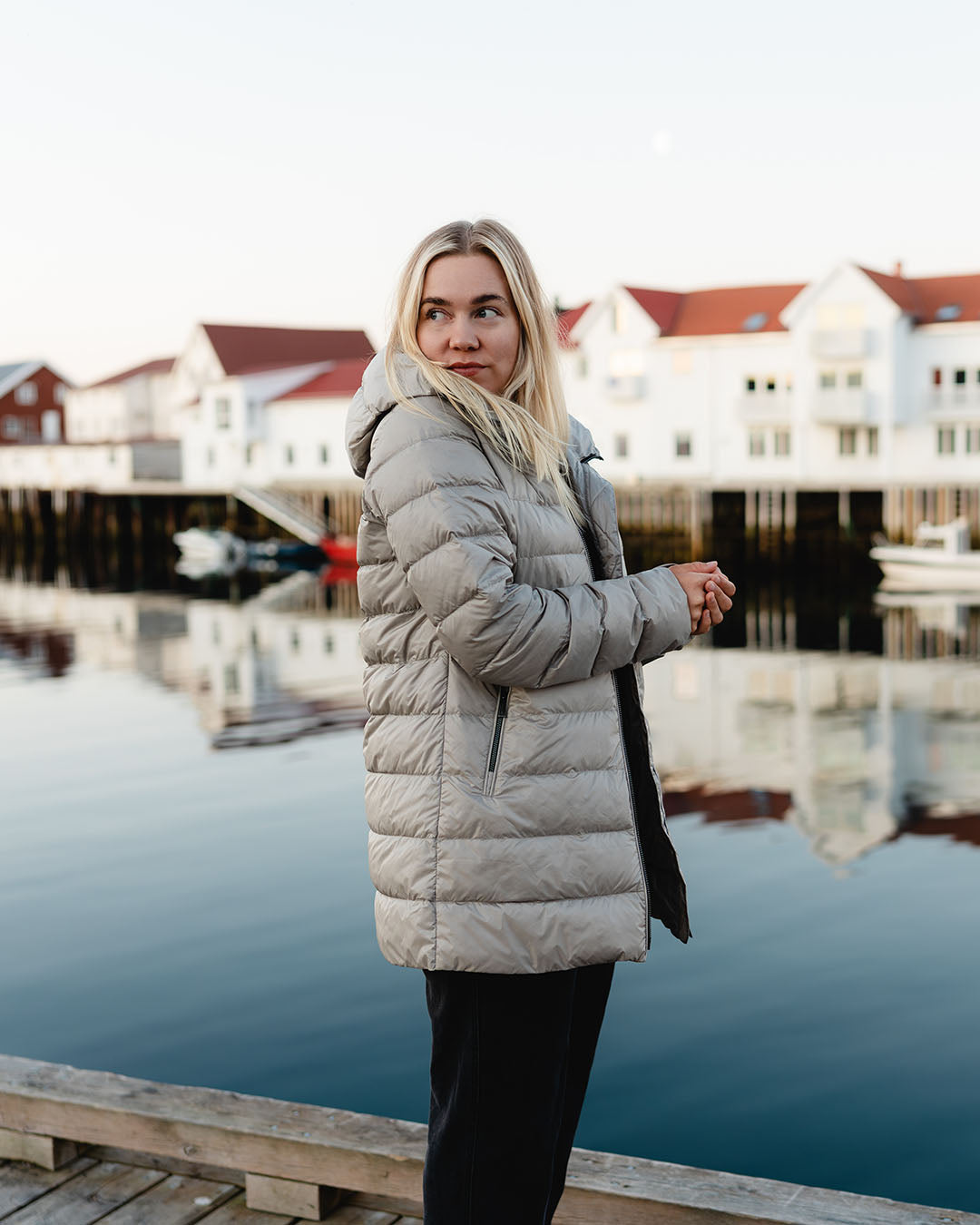 A woman stands on a pier by the water, with light-colored houses and boats of a fishing village in the background. She is wearing a beige Ruut light down coat (color: Champagne) with a hood and zippered pockets.