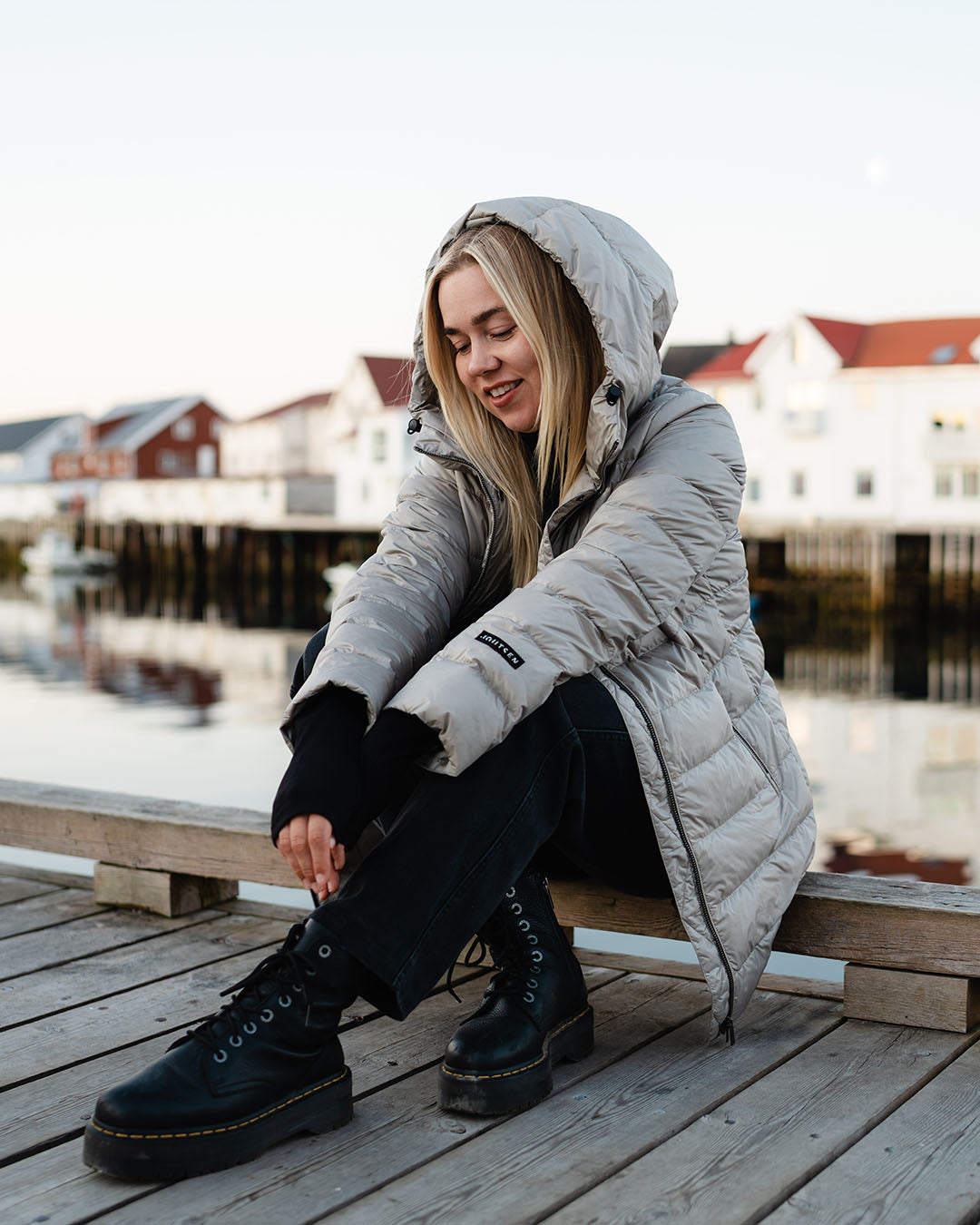 A woman sits on a wooden pier wearing a beige Ruut light down coat (color: champagne) and ties the laces of her black hiking boots. In the background, red-roofed buildings by the water are visible.