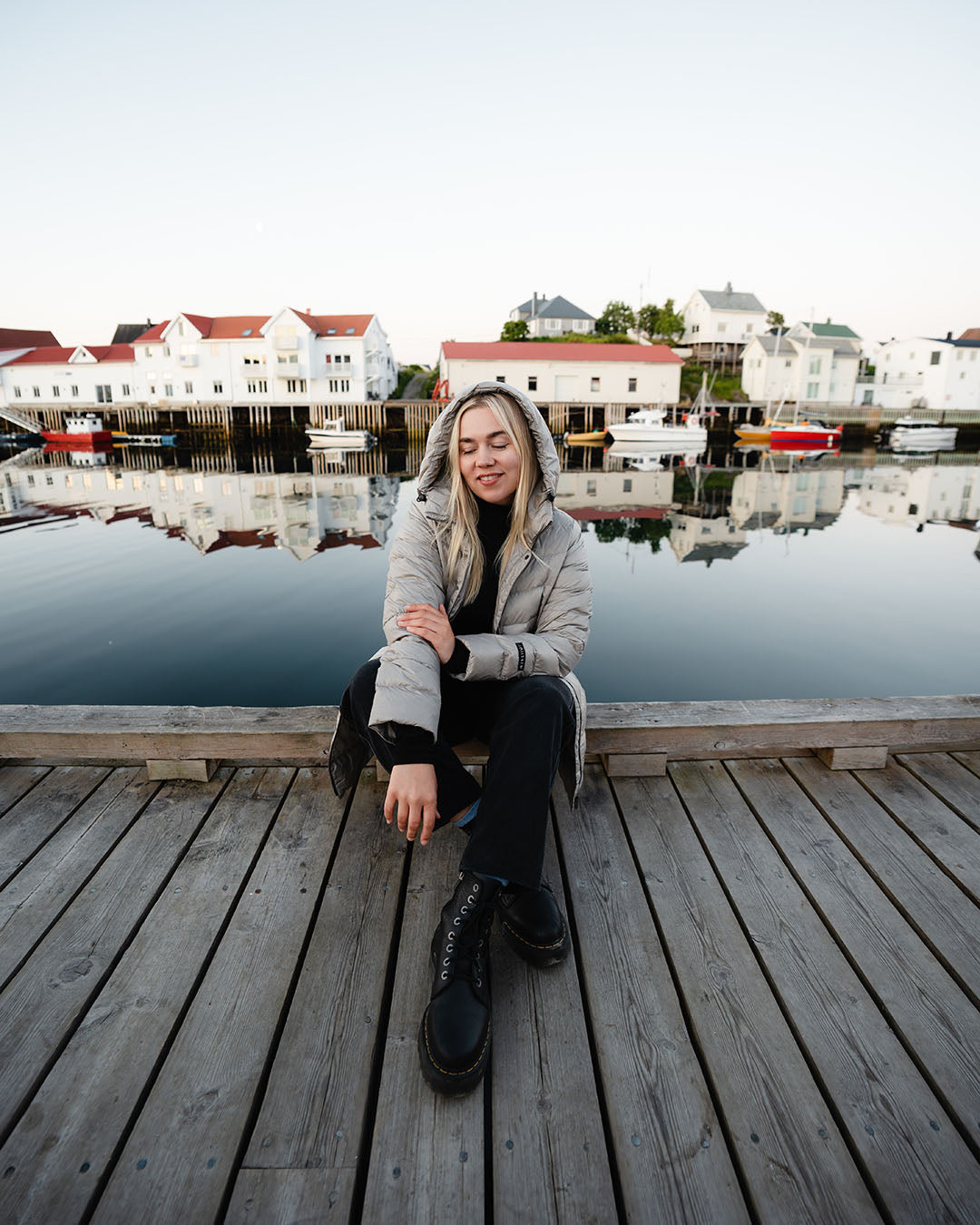 A woman sits on a wooden pier wearing a beige Ruut light down coat (color: champagne). In the background, white buildings and boats by the water are reflected in the calm water.