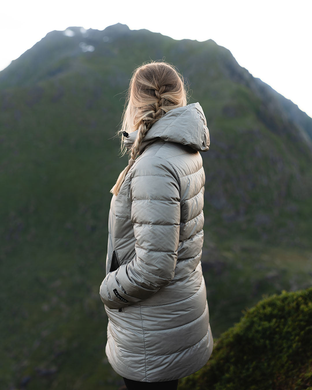 A woman stands in a mountain landscape with her back to the camera, looking out at the view. She is wearing a beige Ruut light down coat (color: Champagne) with a hood. Her hair is casually braided.