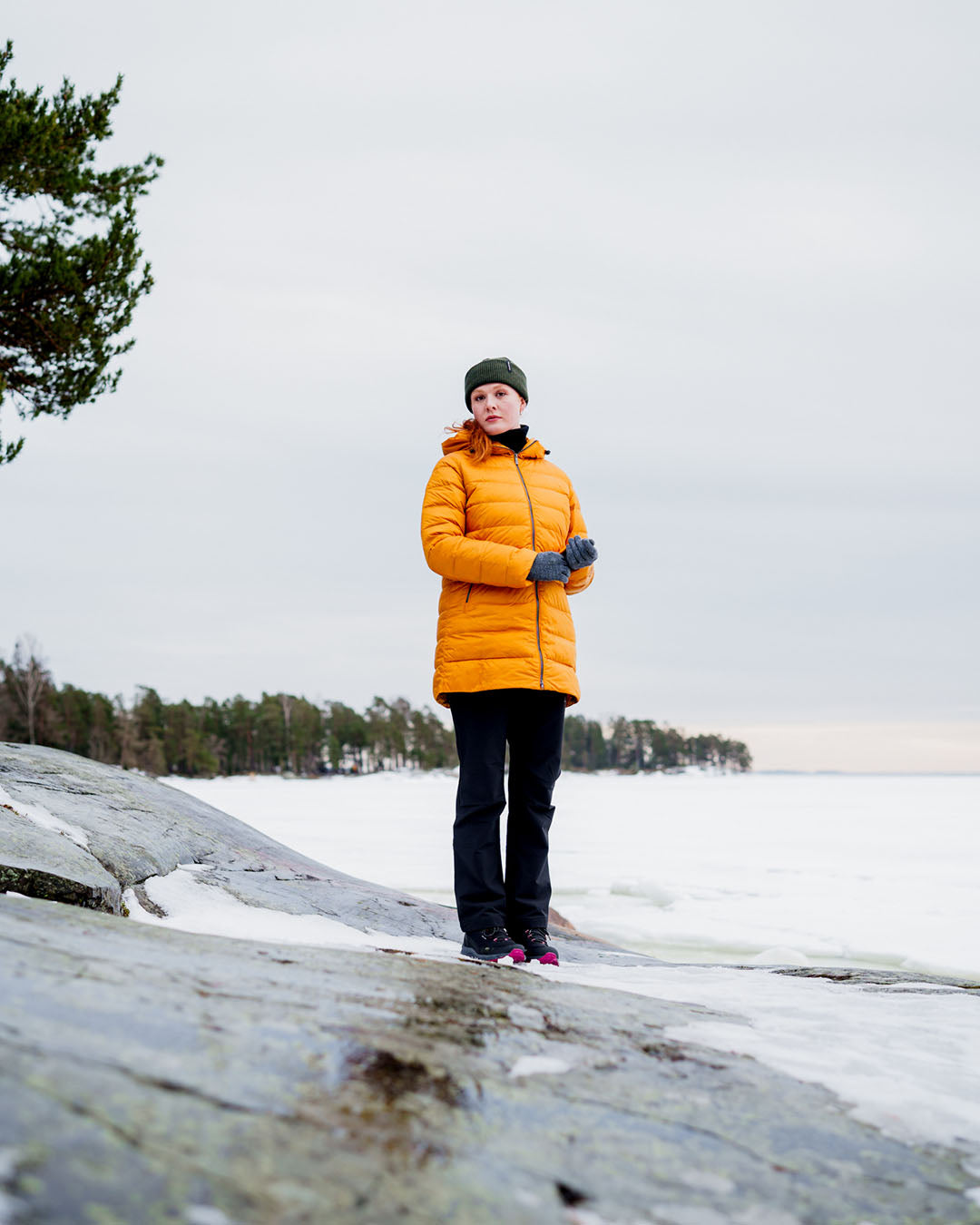 A woman stands on a snowy cliff wearing a yellow Ruut light down coat (color: amber), dark trousers, gray gloves, and a green beanie. In the background, a frozen body of water and a forested shore are visible.
