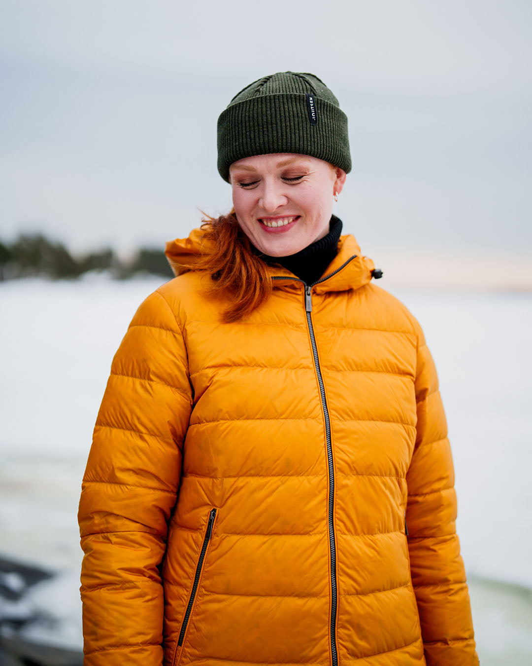 A woman smiles while looking down, wearing a horizontally quilted yellow Ruut light down coat (color: amber) and a green beanie. A snowy landscape is visible in the background.