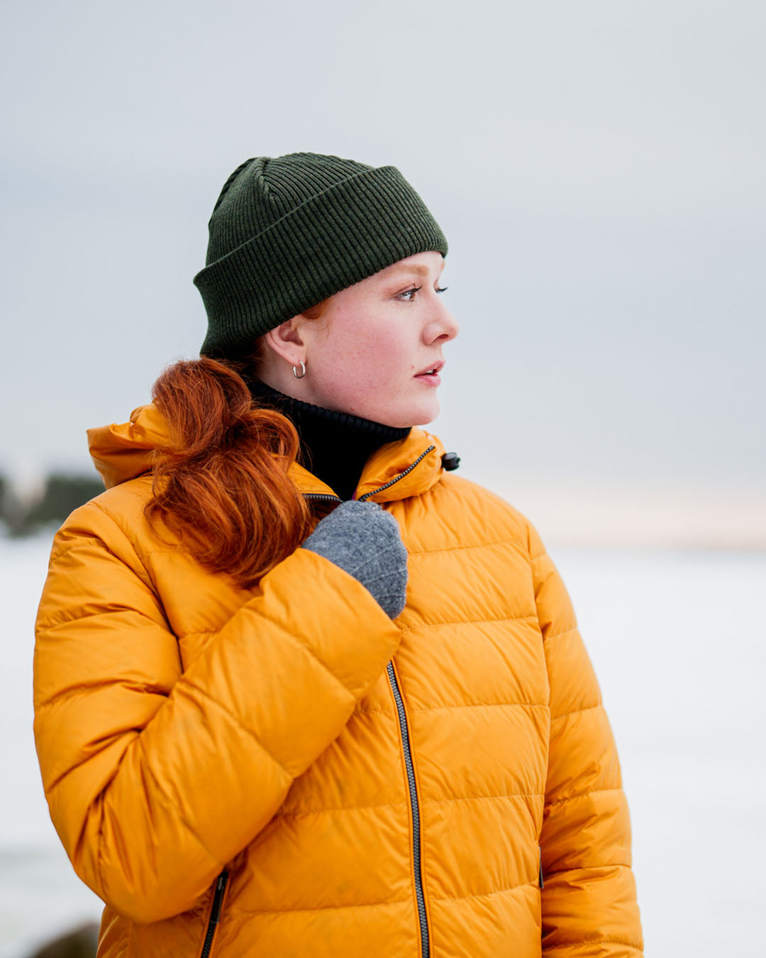 A woman looks to the side, wearing a yellow Ruut light down coat (color: amber), a green beanie, and gray knitted mittens. A snowy landscape is in the background.