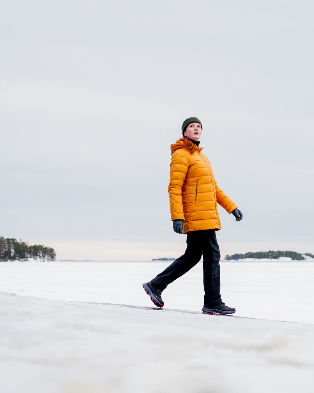 A woman walks on snowy ice, wearing a horizontally quilted yellow Ruut light down coat (color: amber), black trousers, and a green beanie. In the background, there is forest and islands on the horizon.