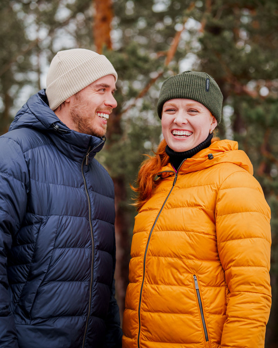 Two people are standing outdoors in a wintry forest landscape. The person on the right is wearing a yellow Ruut light down coat (color: amber) and a green beanie, while the person on the left is wearing a dark blue light down coat and a light-colored beanie. Both are smiling.